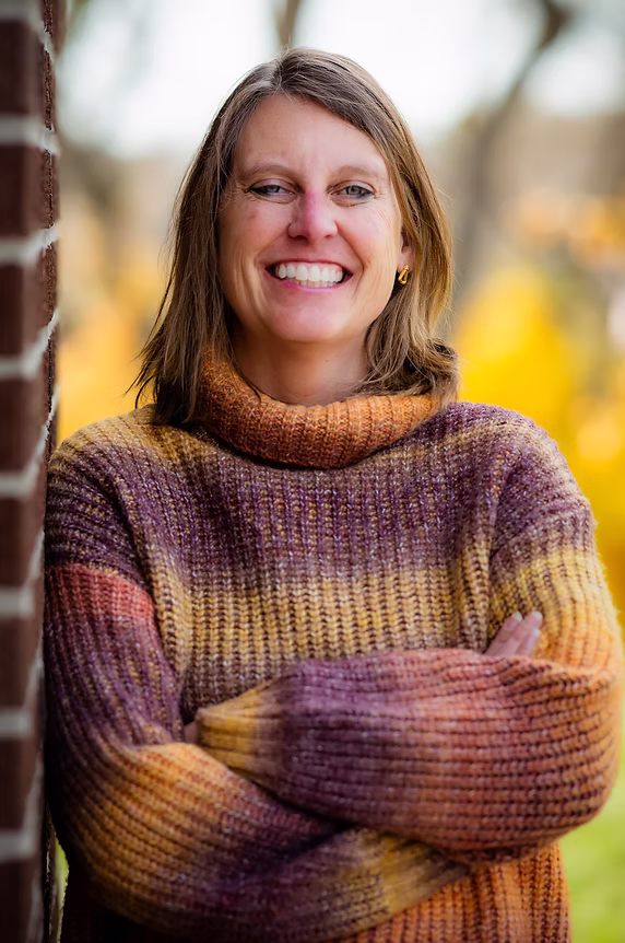 Smiling woman in a striped sweater stands outdoors with arms crossed near a brick wall.