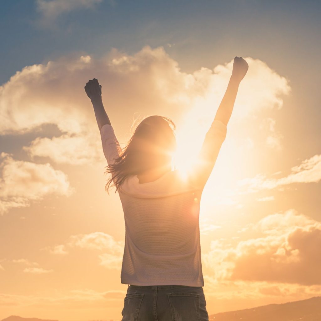 Person standing with arms raised, facing the sunrise or sunset, with sunlight shining through clouds.